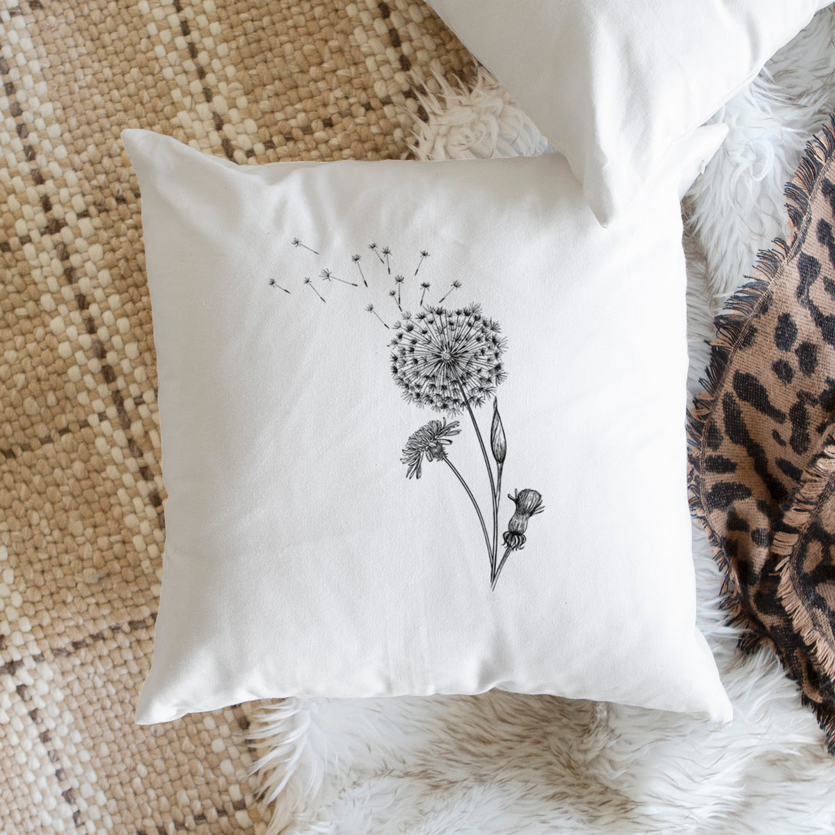 White pillow cover printed with a black ink line drawing of a dandelion seed head with seeds blowing away, on a woven chair.