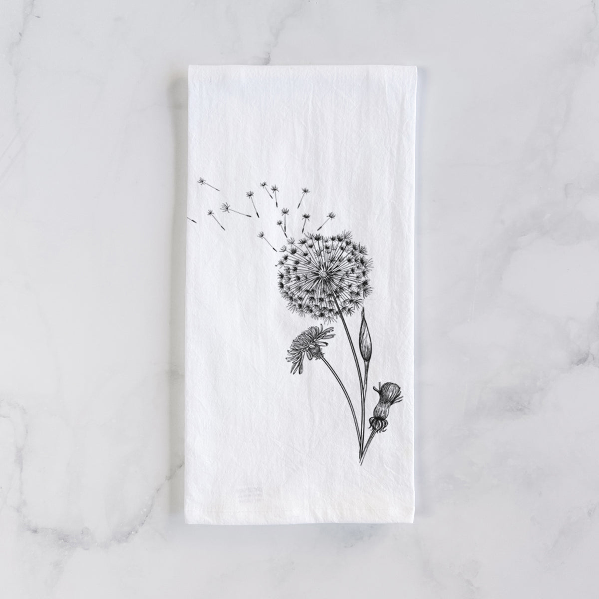 White cotton tea towel laid flat on marble, printed with a black ink line drawing of a dandelion seed head with seeds blowing away.