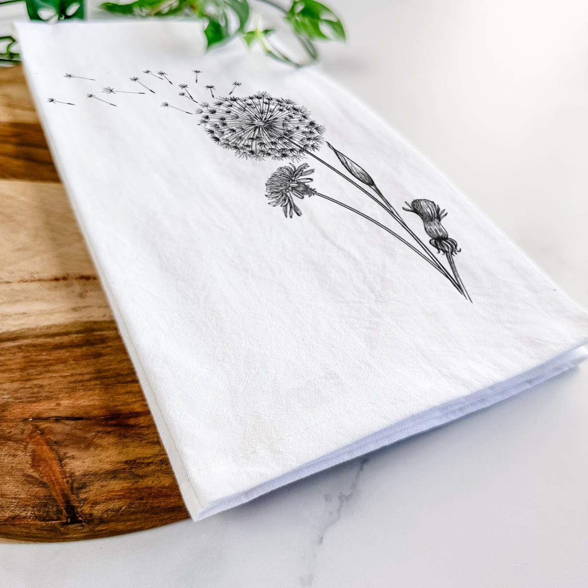Close-up of a folded white tea towel on a wooden board, showing a black ink line drawing of a dandelion seed head with seeds blowing away.