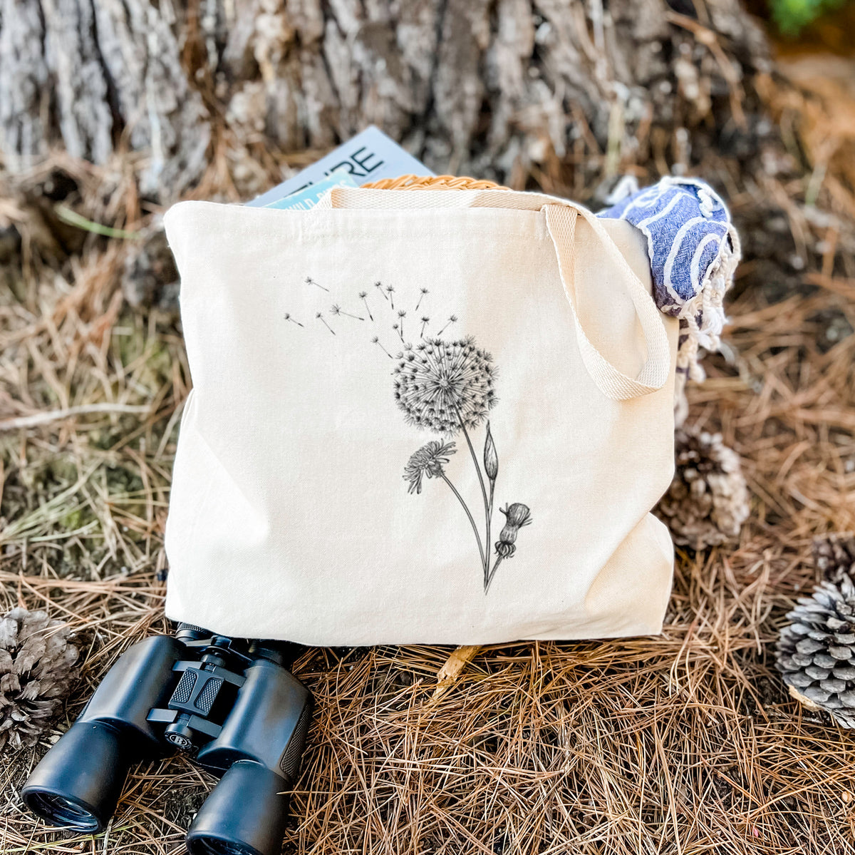 Canvas tote bag printed with a black ink line drawing of a dandelion seed head with seeds blowing away, resting on dry grass beside a pair of binoculars.