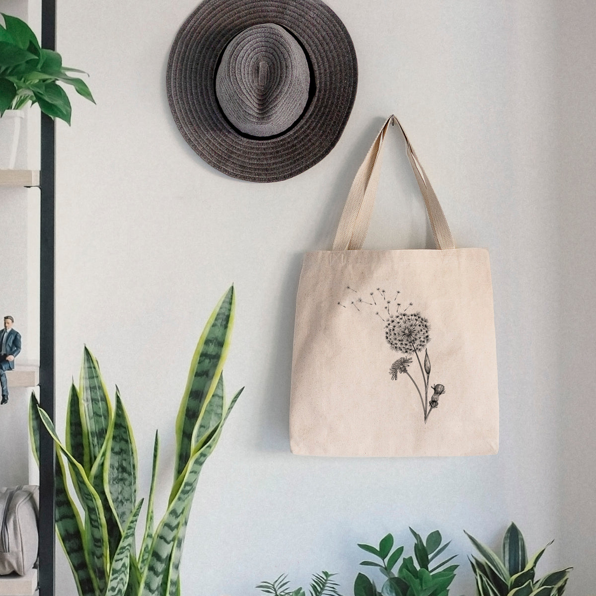 Canvas tote bag printed with a black ink line drawing of a dandelion seed head with seeds blowing away, hanging on a wall hook below a hat, next to potted plants.