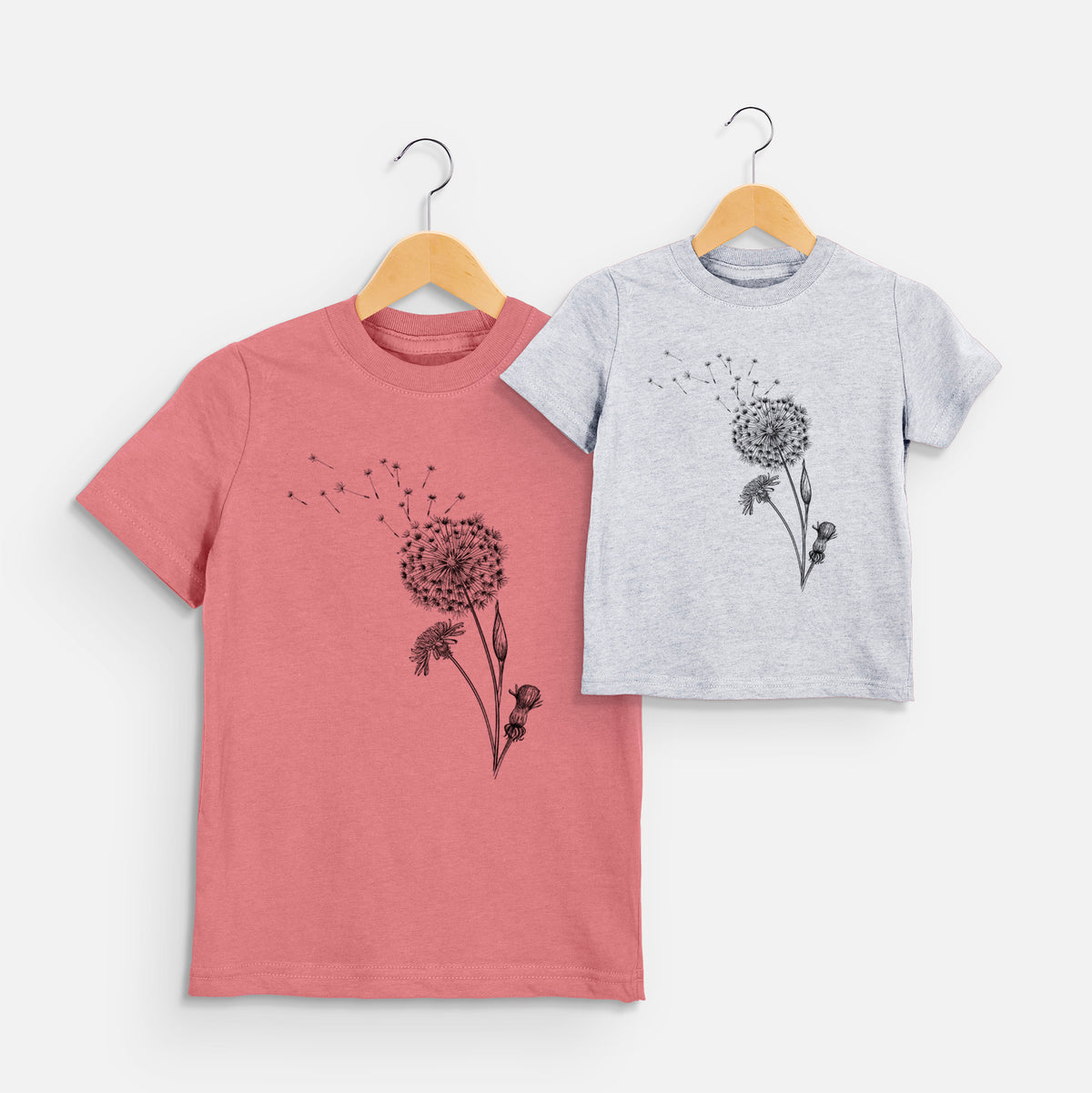 Two youth t-shirts in different colors on hangers, printed with a black ink line drawing of a dandelion seed head with seeds blowing away.