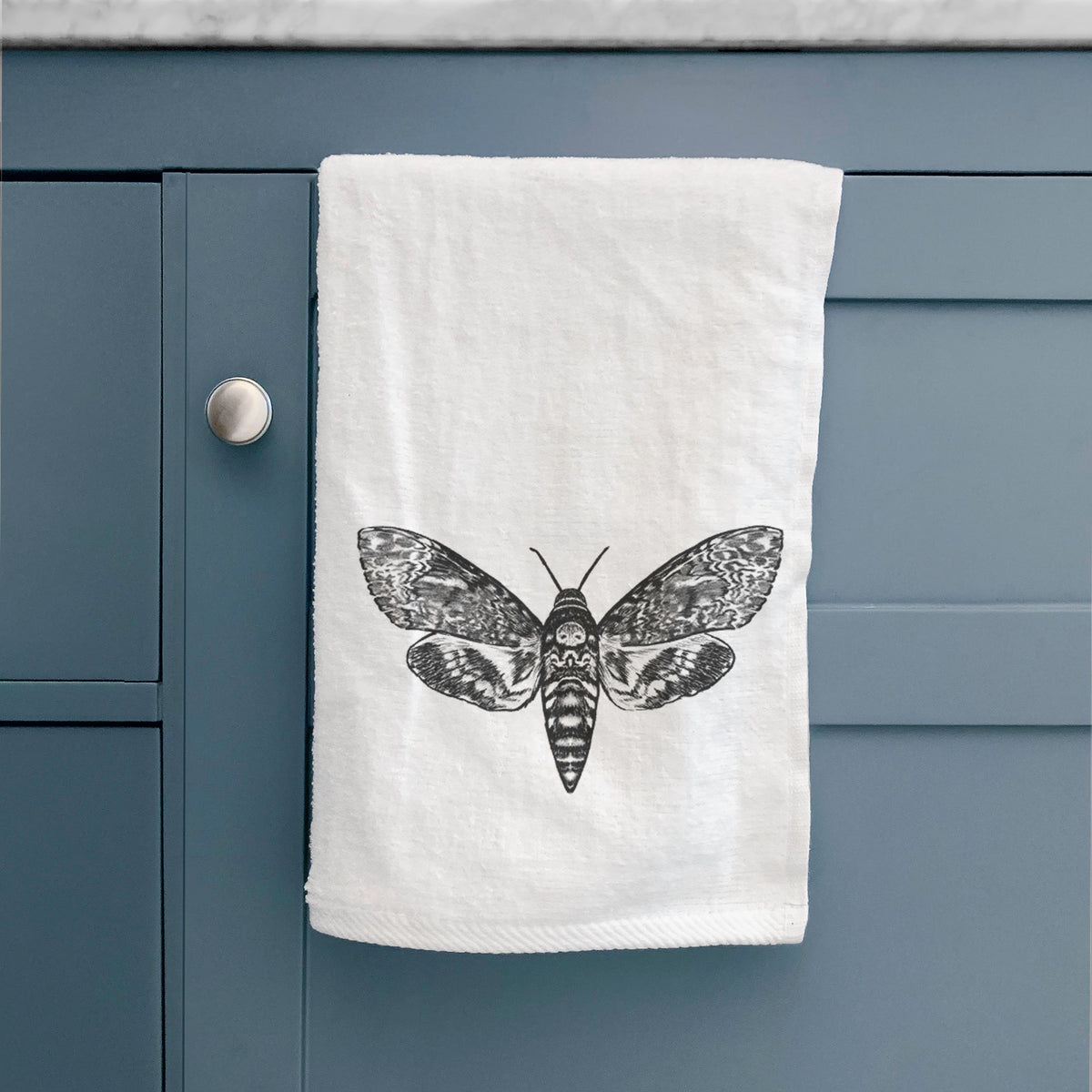 White hand towel printed with a detailed black ink illustration of a death's-head hawkmoth with its wings spread, hanging on a cabinet door.