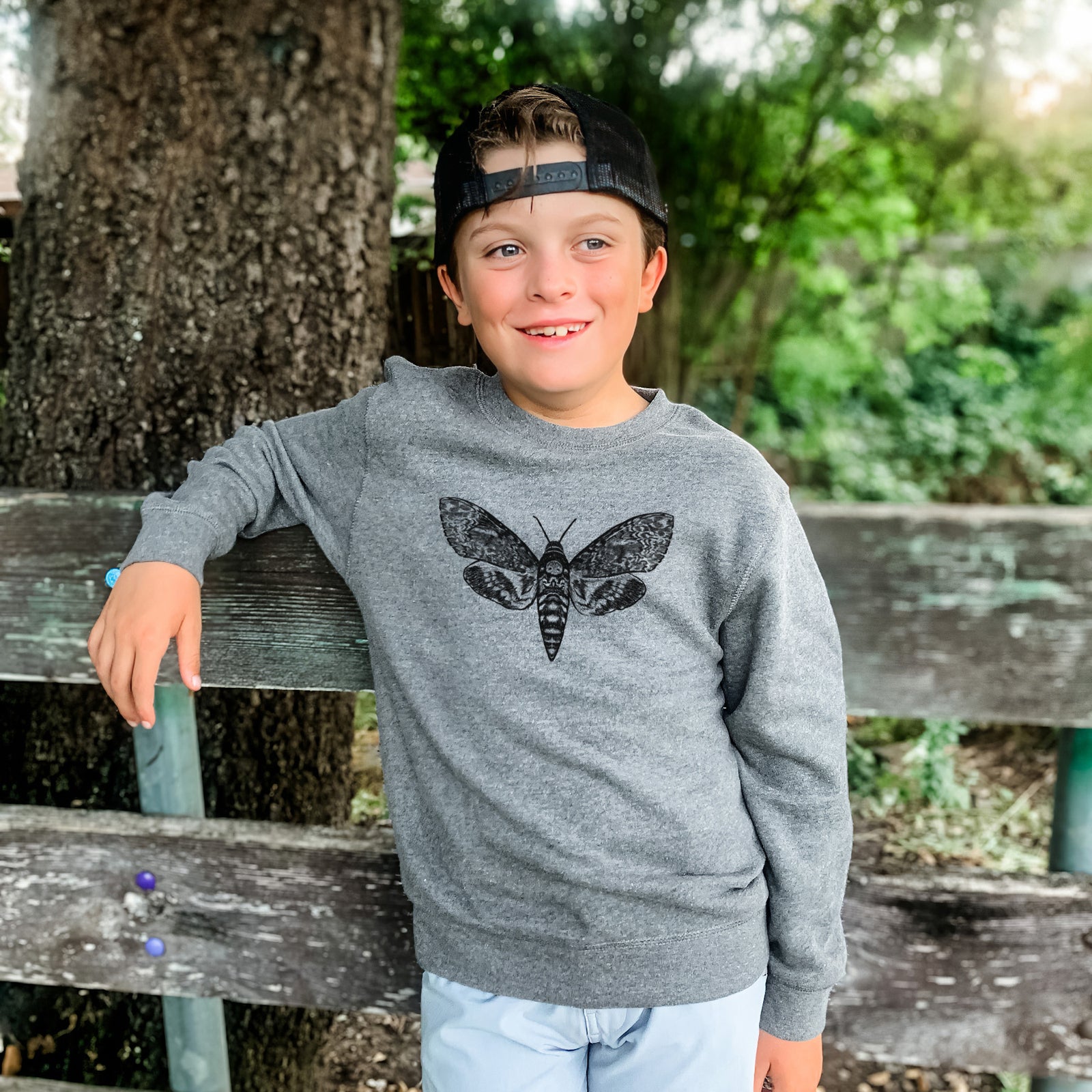 Boy wearing a youth crewneck sweatshirt printed with a detailed black ink illustration of a death's-head hawkmoth with its wings spread, outdoors.
