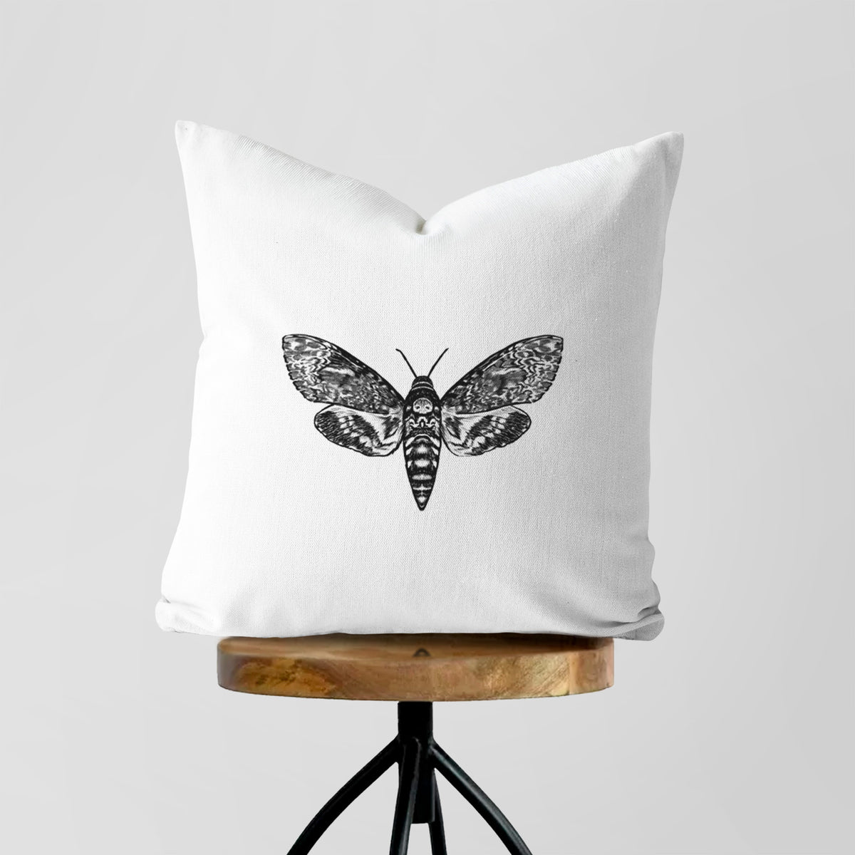 White pillow cover printed with a detailed black ink illustration of a death's-head hawkmoth with its wings spread, on a wooden stool.