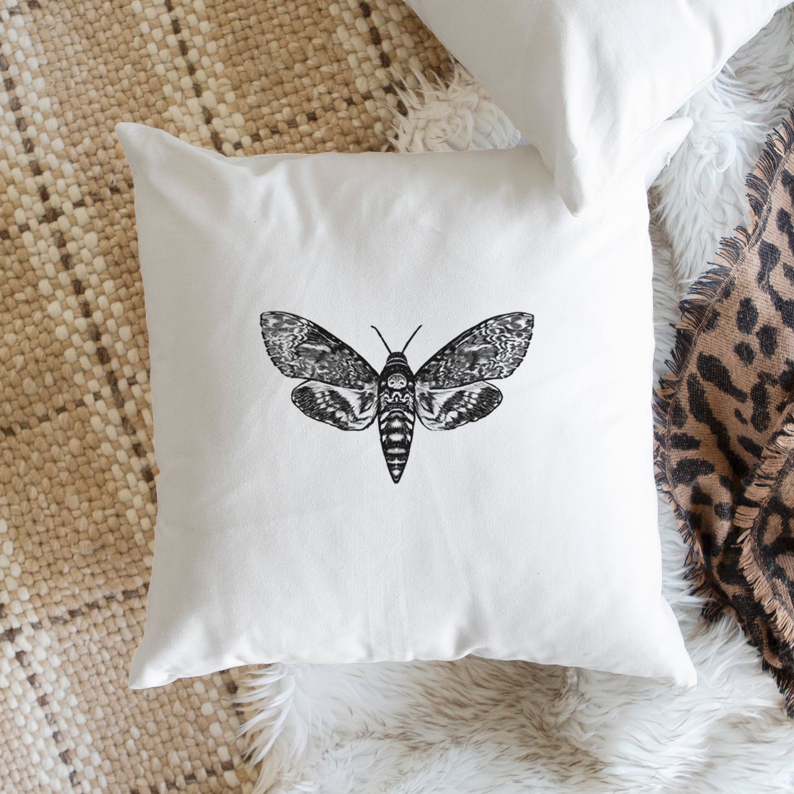 White pillow cover printed with a detailed black ink illustration of a death's-head hawkmoth with its wings spread, on a bench beside plants.