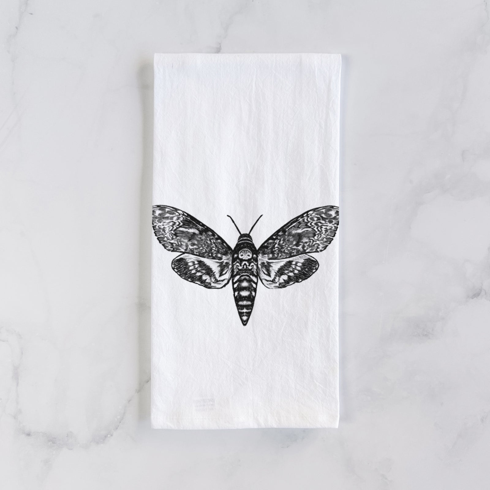 White cotton tea towel printed with a detailed black ink illustration of a death's-head hawkmoth with its wings spread, on a marble counter beside a round wooden cutting board and greenery.