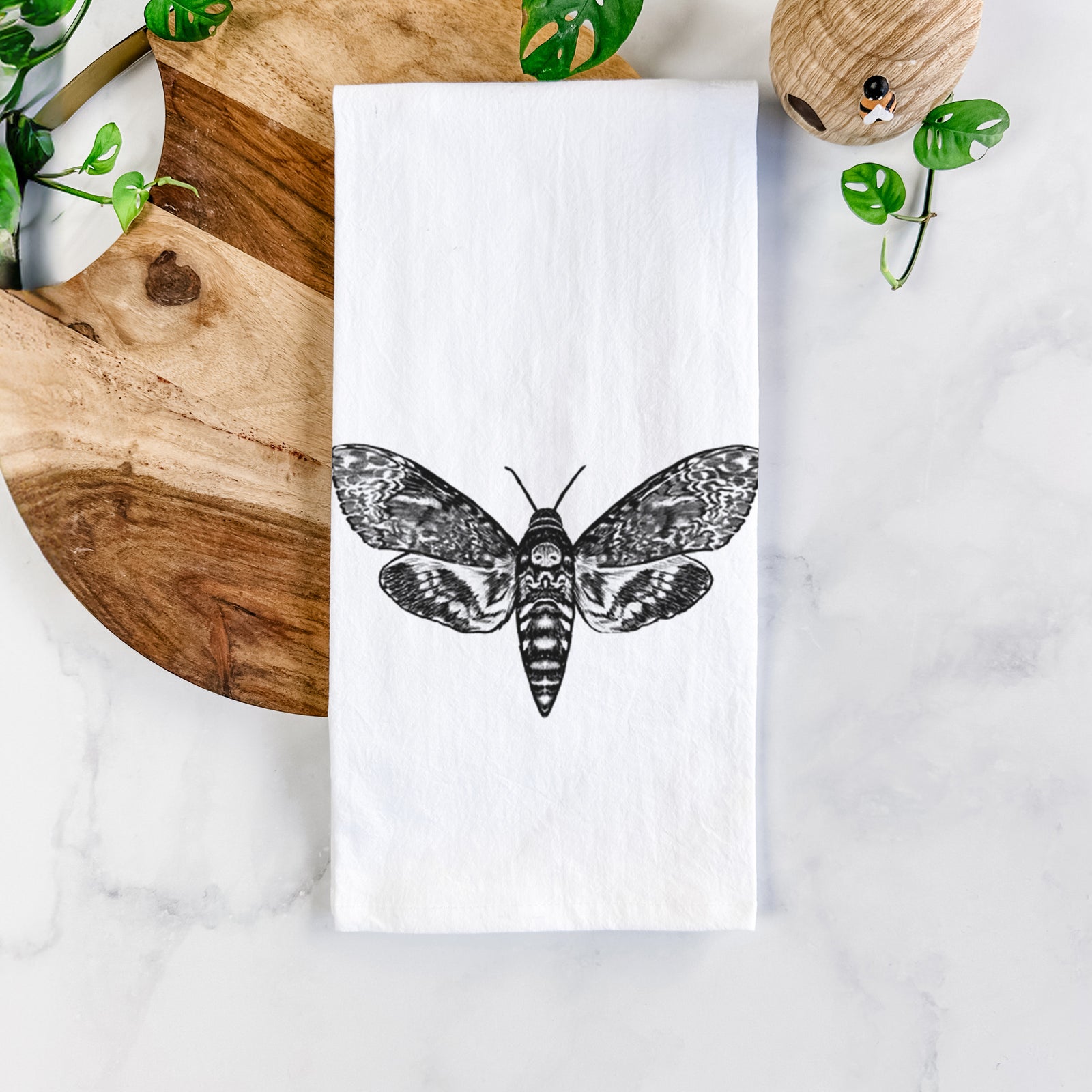 White cotton tea towel printed with a detailed black ink illustration of a death's-head hawkmoth with its wings spread, on a marble counter beside a round wooden cutting board and greenery.