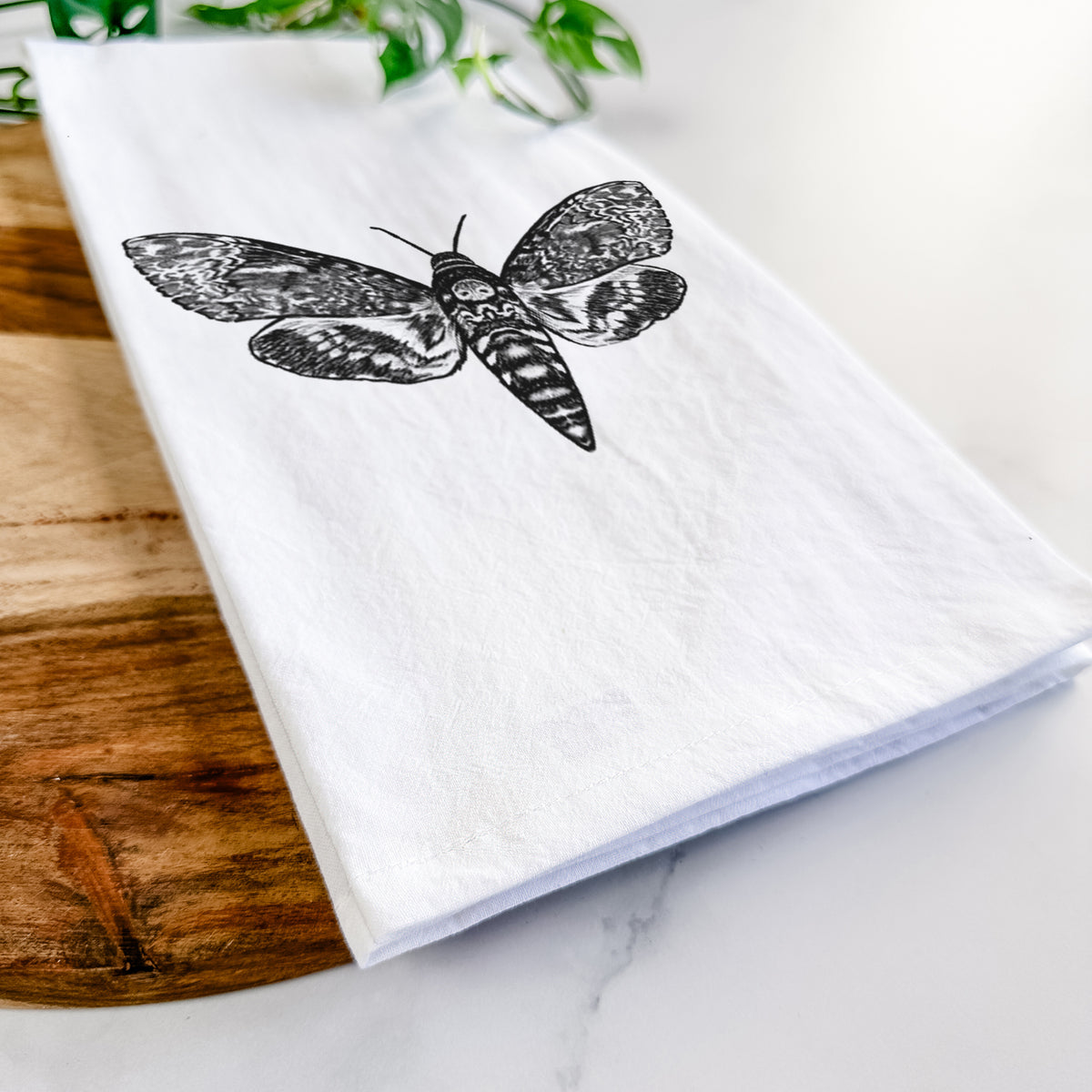 Close-up of a folded white tea towel on a wooden board, showing a detailed black ink illustration of a death's-head hawkmoth with its wings spread.