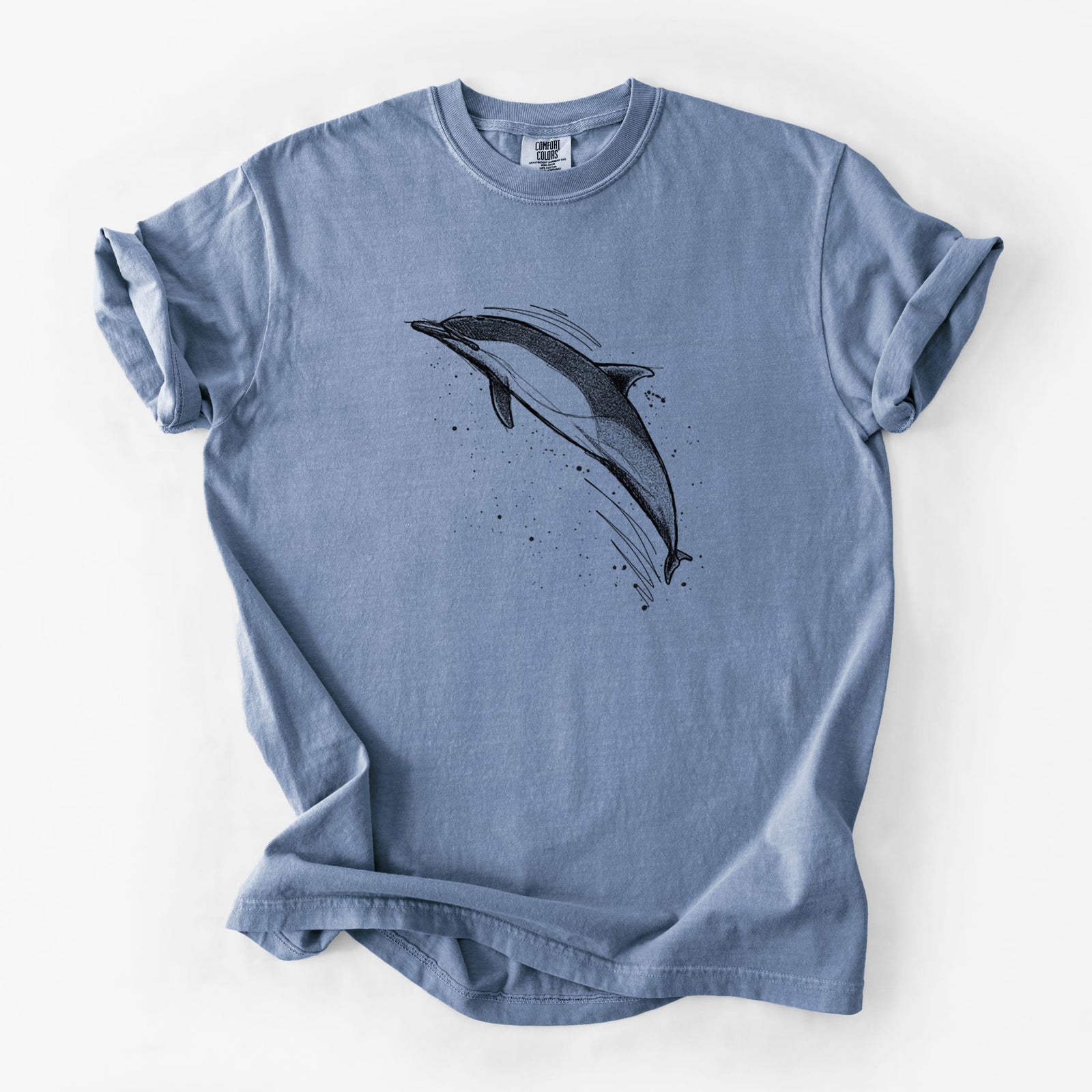 Blue jean Comfort Colors t-shirt laid flat, printed with a black ink illustration of a common dolphin leaping, with ink splatters.