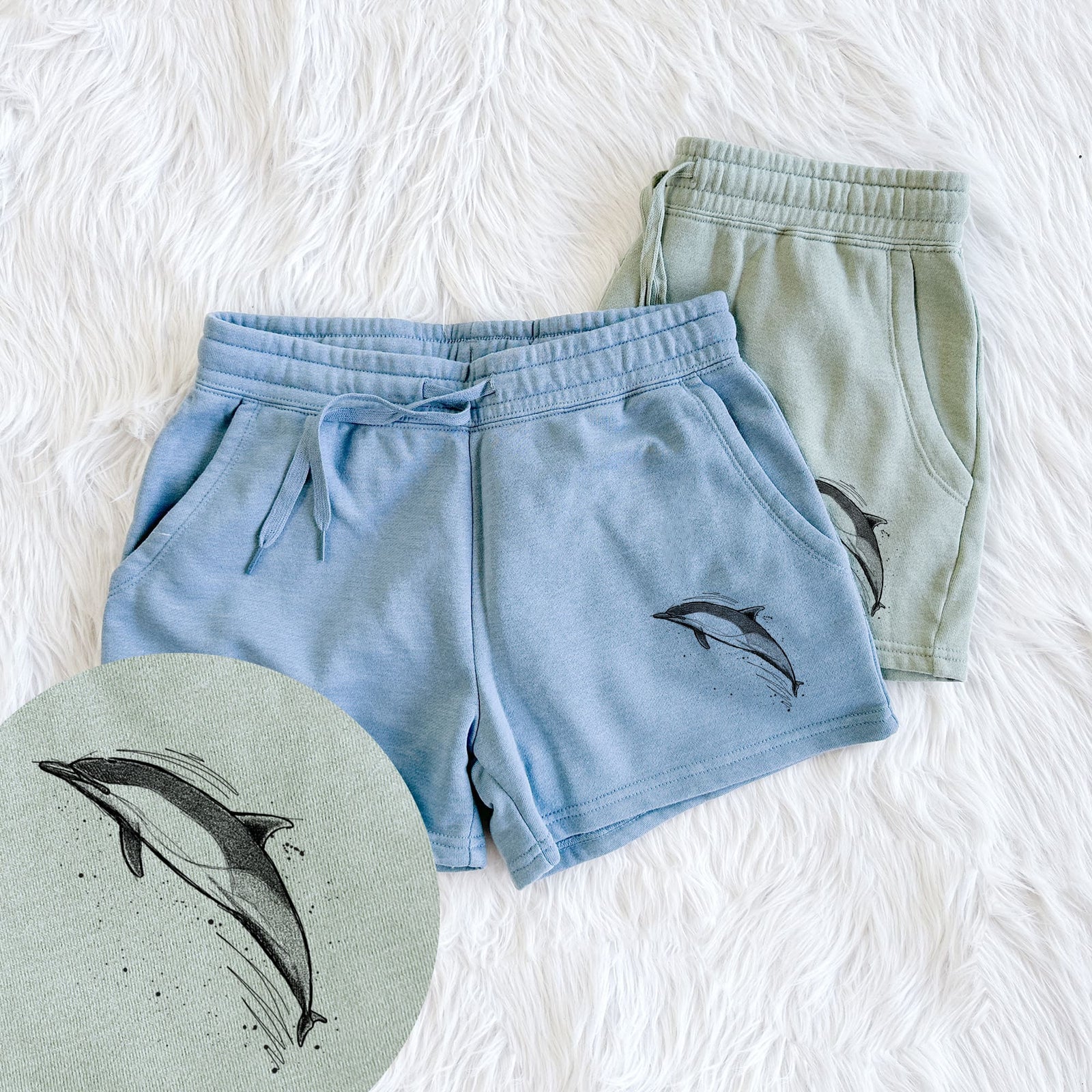 Two pairs of Cali Wave shorts in different colors printed with a black ink illustration of a common dolphin leaping, with ink splatters, with a close-up of the print.
