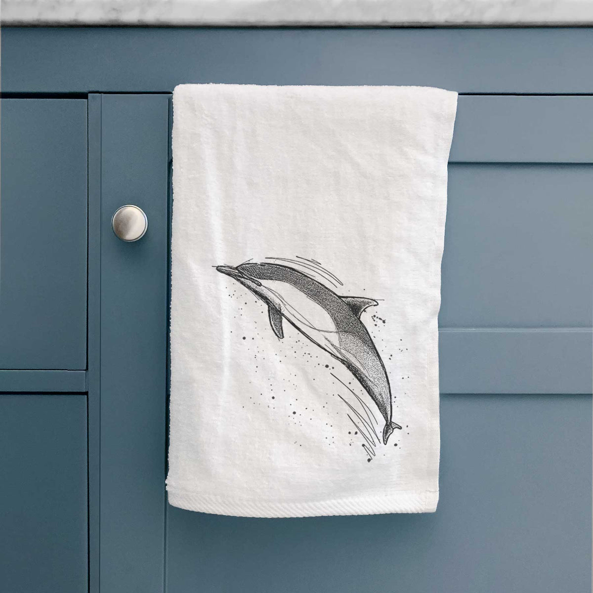 White hand towel printed with a black ink illustration of a common dolphin leaping, with ink splatters, hanging on a cabinet door.