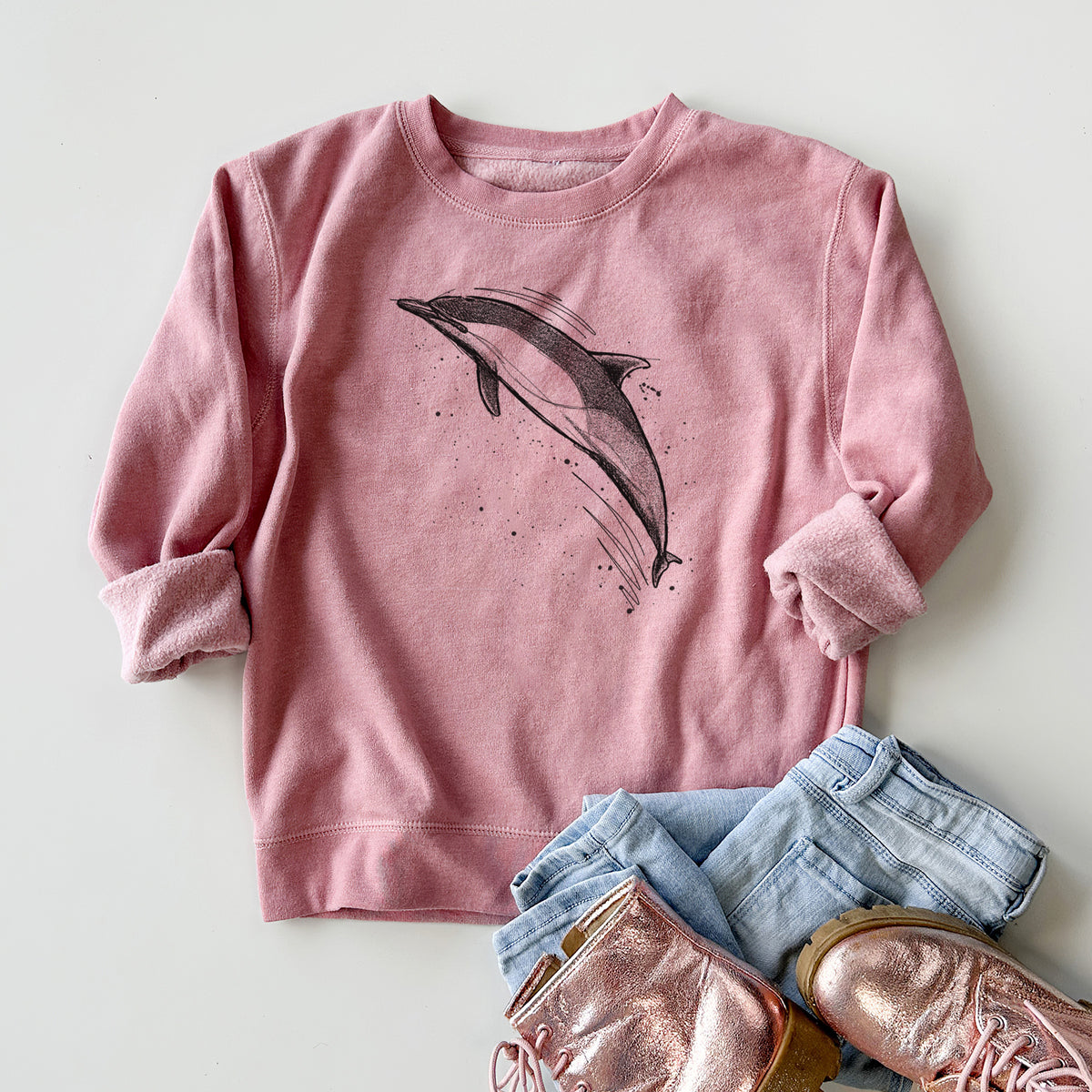 Youth crewneck sweatshirt printed with a black ink illustration of a common dolphin leaping, with ink splatters, styled flat with jeans and boots.