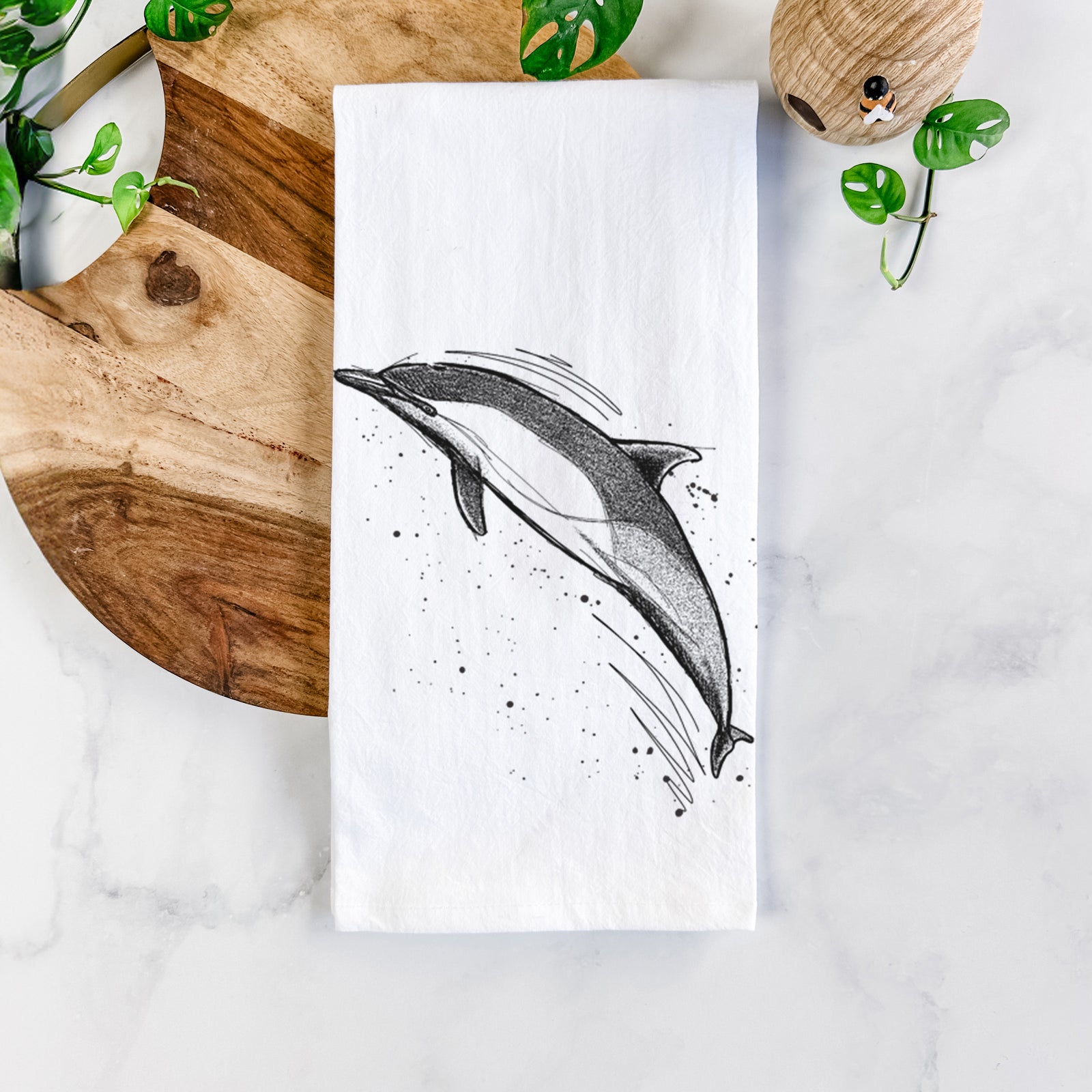 White cotton tea towel printed with a black ink illustration of a common dolphin leaping, with ink splatters, on a marble counter beside a round wooden cutting board and greenery.
