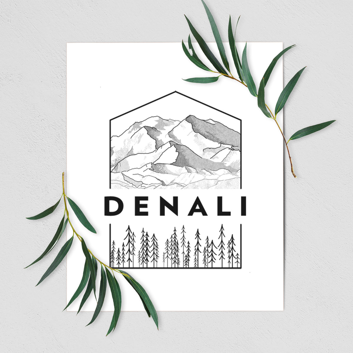 Art print of a black line art badge of the Denali mountain range above pine trees, with the word Denali, shown with eucalyptus leaves.