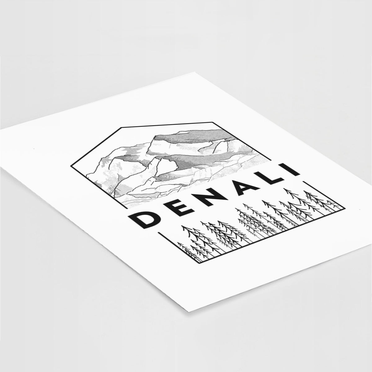 Art print of a black line art badge of the Denali mountain range above pine trees, with the word Denali, lying at an angle on a white surface.