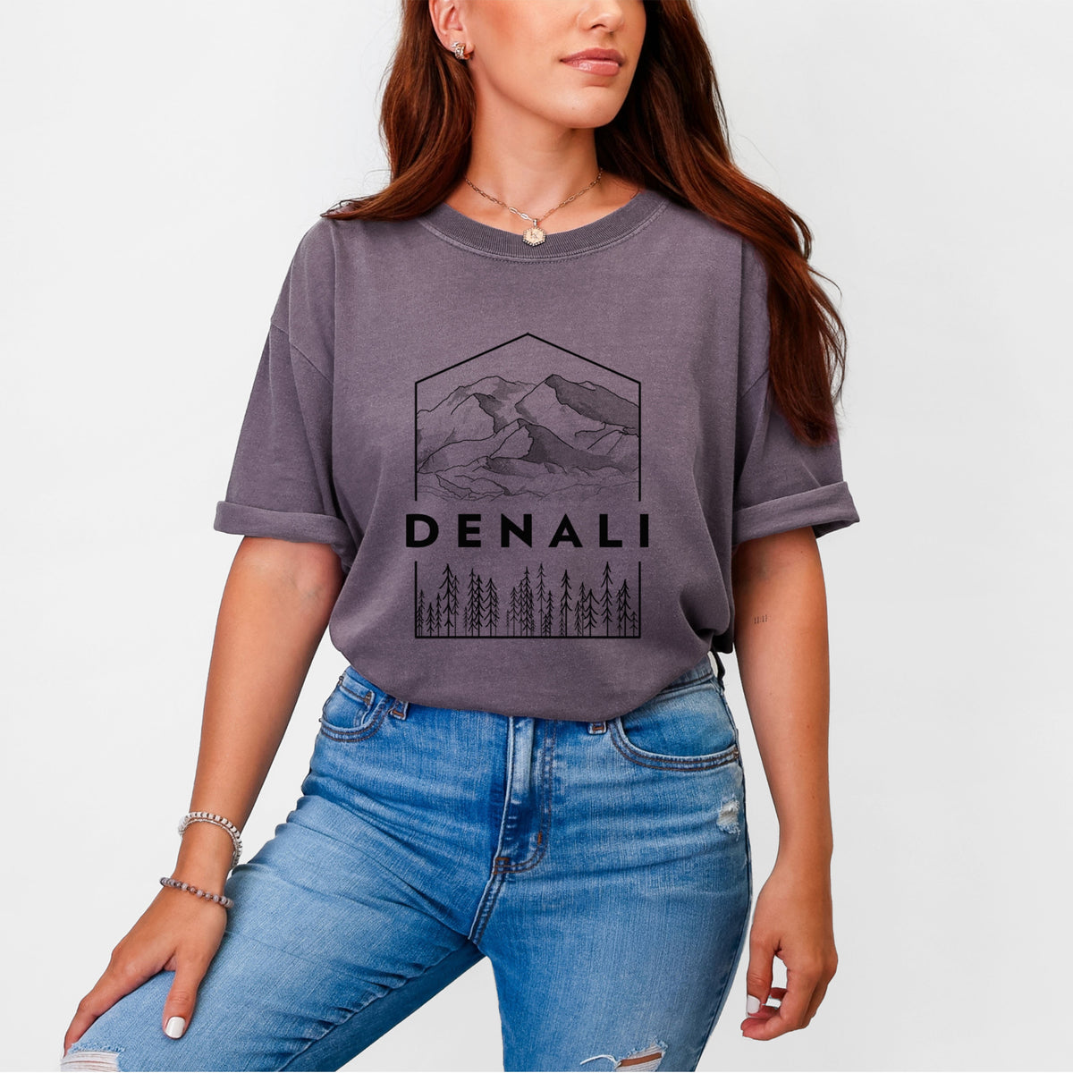 Woman wearing a Comfort Colors t-shirt printed with a black line art badge of the Denali mountain range above pine trees, with the word Denali.