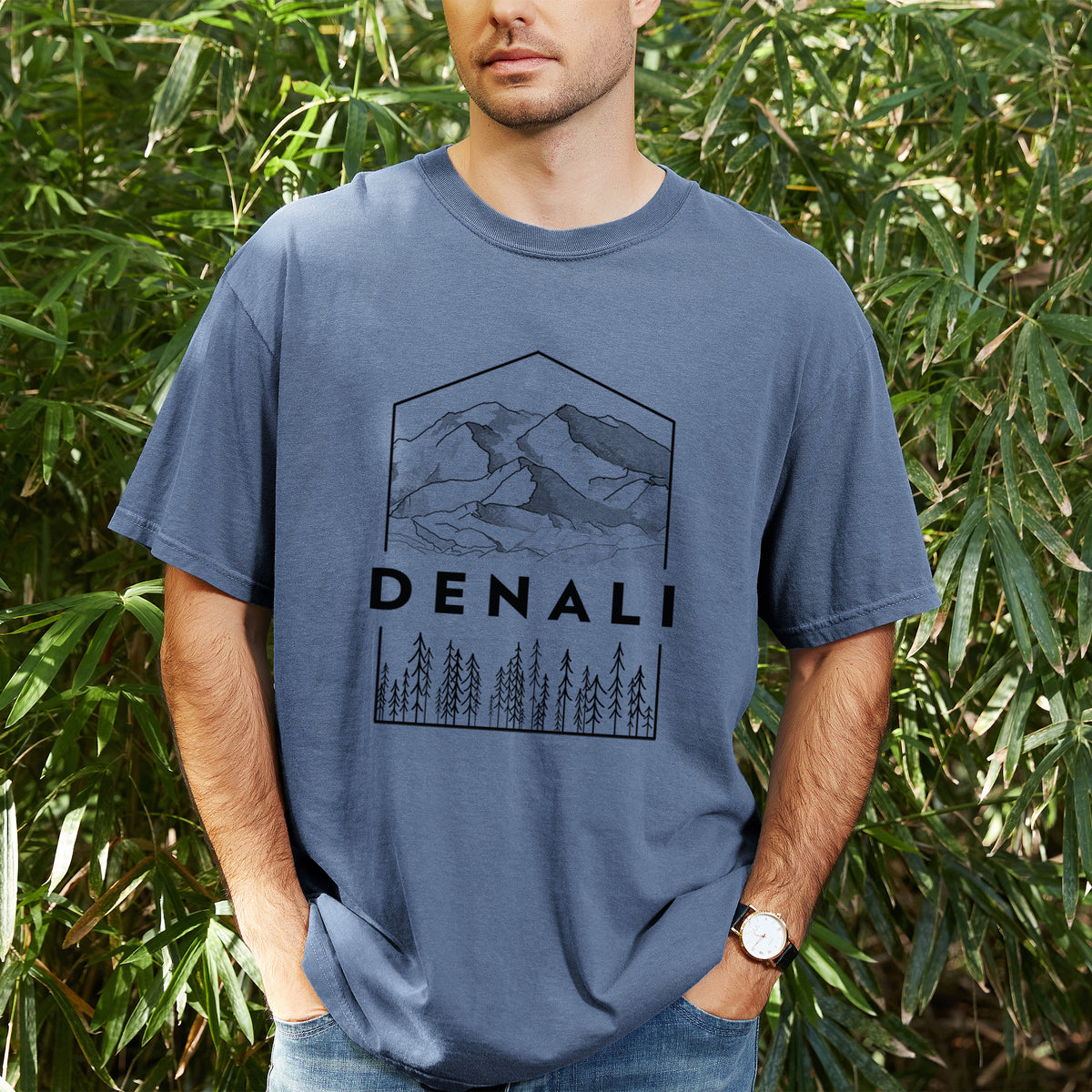 Man wearing a Comfort Colors t-shirt printed with a black line art badge of the Denali mountain range above pine trees, with the word Denali, outdoors.