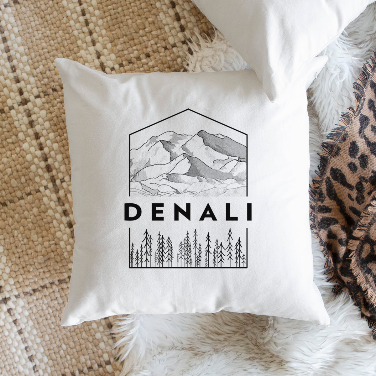 White pillow cover printed with a black line art badge of the Denali mountain range above pine trees, with the word Denali, on a woven chair.