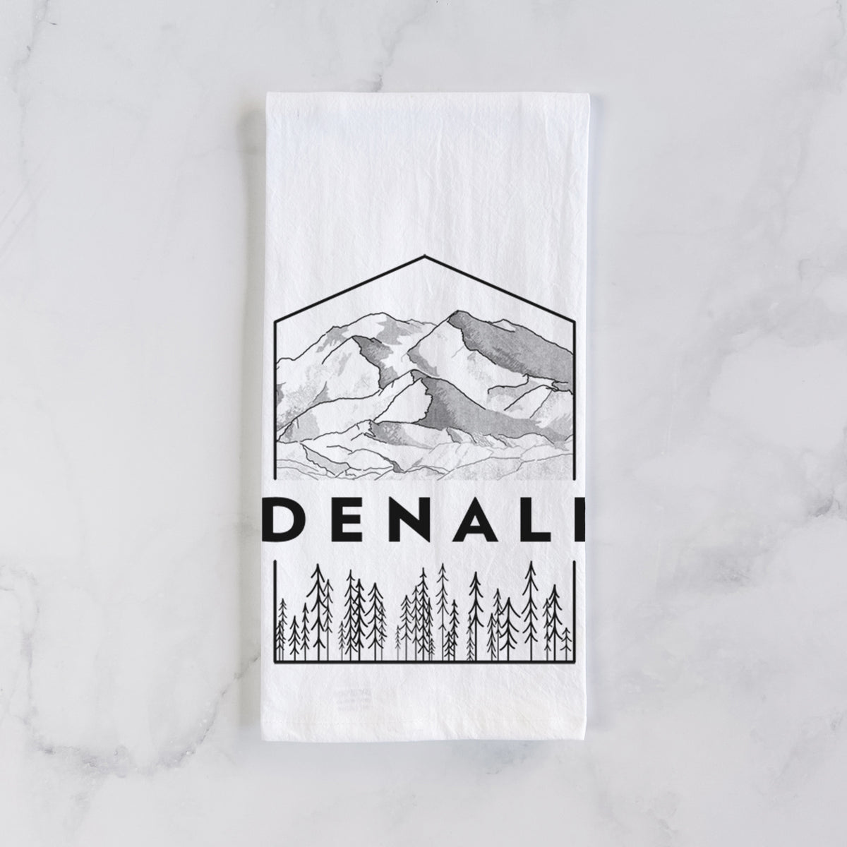 White cotton tea towel laid flat on marble, printed with a black line art badge of the Denali mountain range above pine trees, with the word Denali.