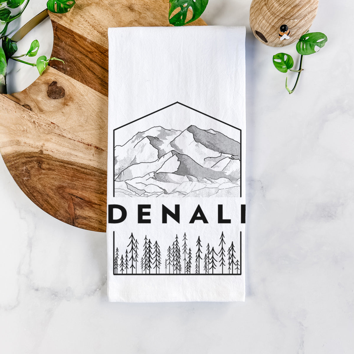 White cotton tea towel printed with a black line art badge of the Denali mountain range above pine trees, with the word Denali, on a marble counter beside a round wooden cutting board and greenery.