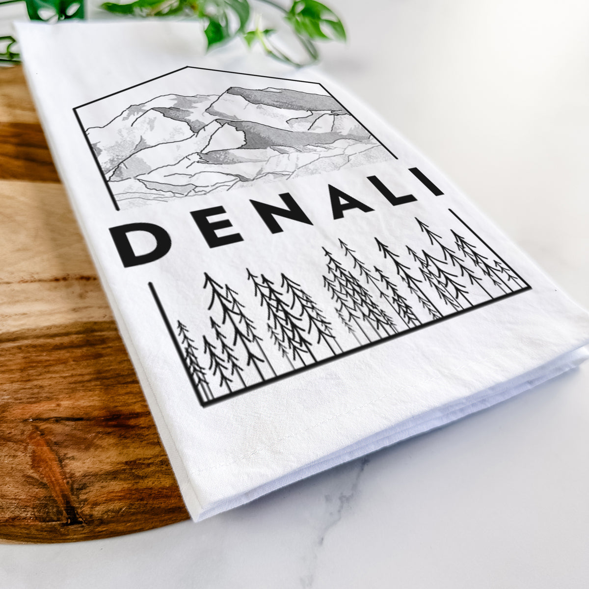 Close-up of a folded white tea towel on a wooden board, showing a black line art badge of the Denali mountain range above pine trees, with the word Denali.