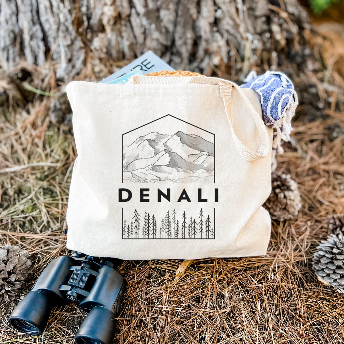 Canvas tote bag printed with a black line art badge of the Denali mountain range above pine trees, with the word Denali, resting on dry grass beside a pair of binoculars.