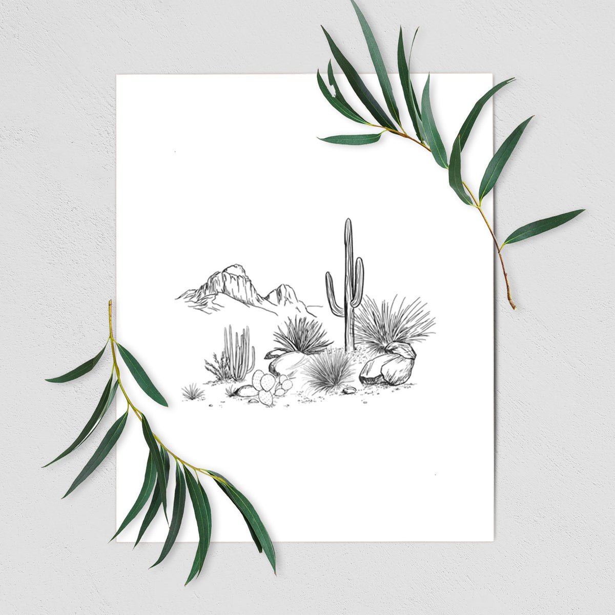 Art print of a black ink desert scene with a saguaro cactus, agave, rocks and distant mountains, shown with eucalyptus leaves.