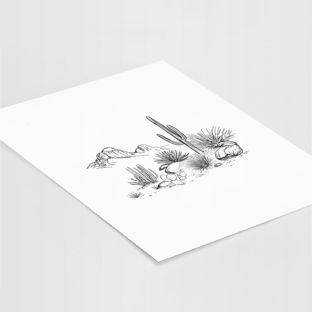 Art print of a black ink desert scene with a saguaro cactus, agave, rocks and distant mountains, lying at an angle on a white surface.