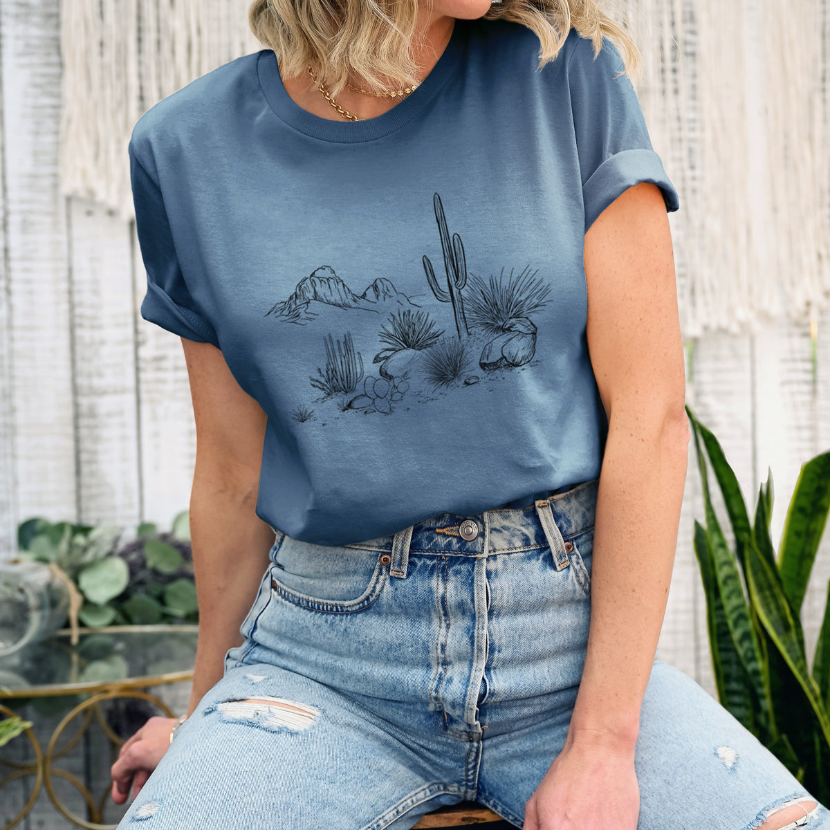 Woman wearing a steel blue lightweight cotton t-shirt printed with a black ink desert scene with a saguaro cactus, agave, rocks and distant mountains.