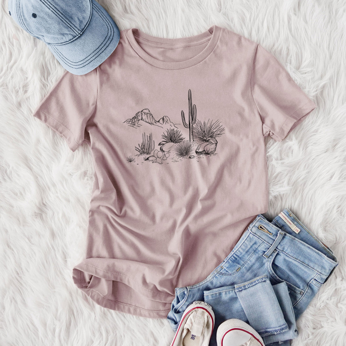 Women's relaxed fit cotton t-shirt printed with a black ink desert scene with a saguaro cactus, agave, rocks and distant mountains, styled flat on a white faux fur rug with jeans.