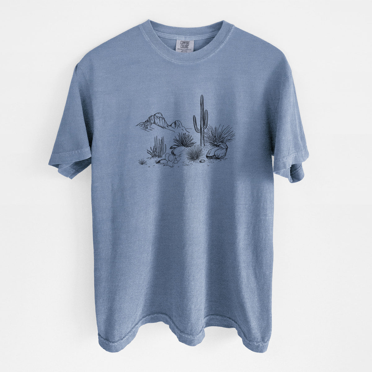 Blue jean Comfort Colors t-shirt laid flat, printed with a black ink desert scene with a saguaro cactus, agave, rocks and distant mountains.
