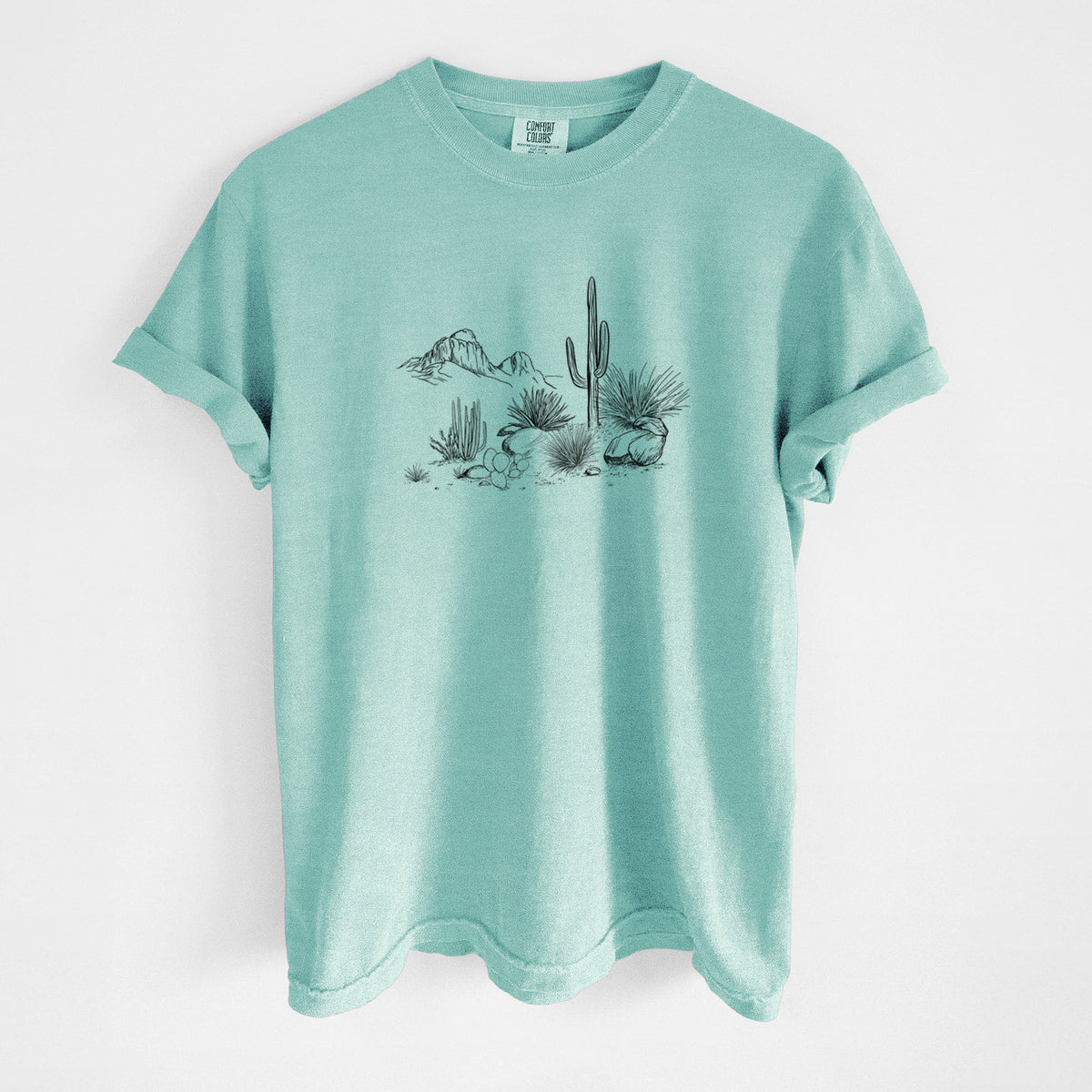 Chalky mint Comfort Colors t-shirt laid flat, printed with a black ink desert scene with a saguaro cactus, agave, rocks and distant mountains.