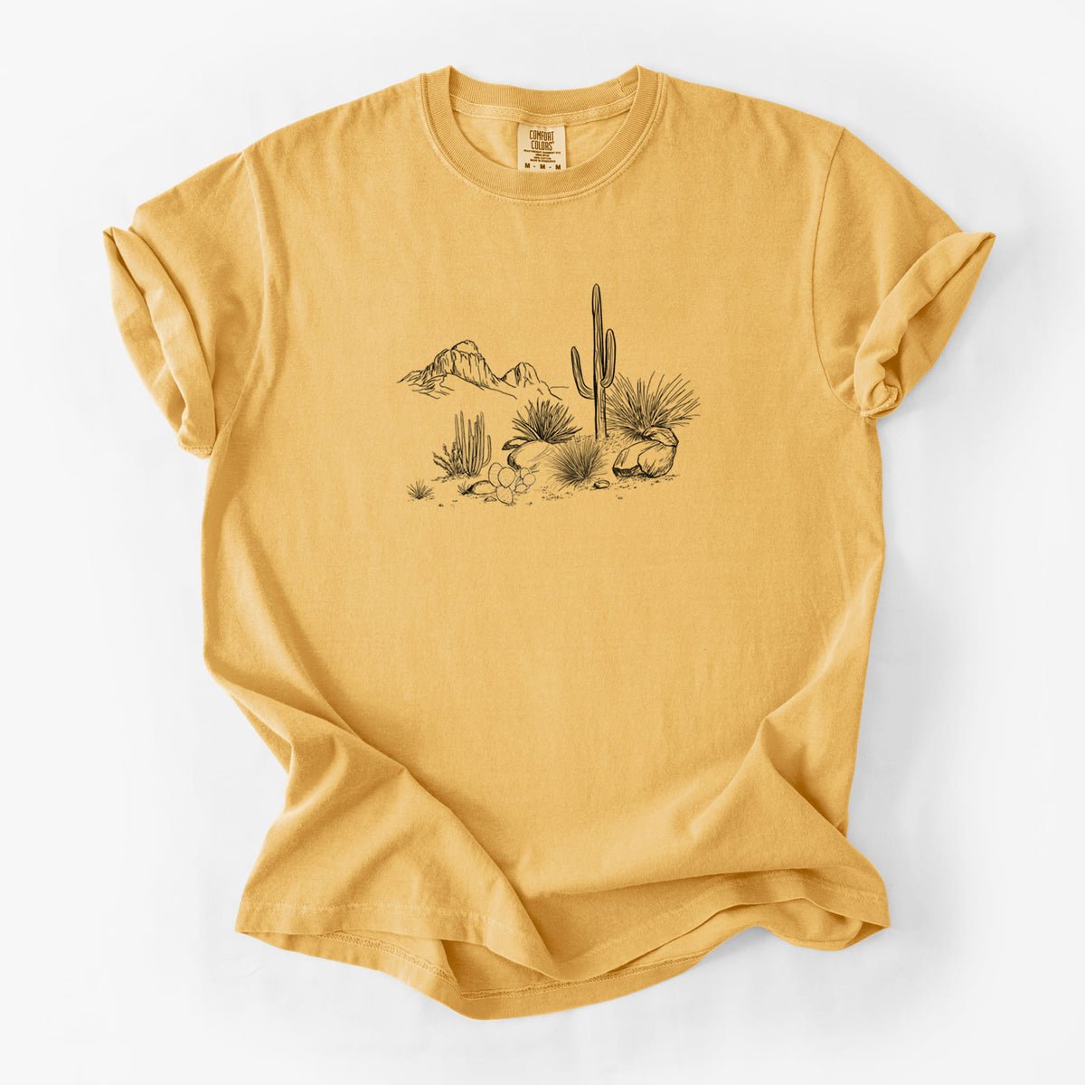 Golden wheat yellow Comfort Colors t-shirt laid flat, printed with a black ink desert scene with a saguaro cactus, agave, rocks and distant mountains.