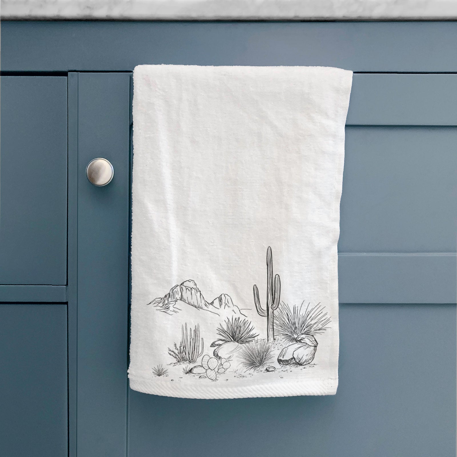 White hand towel printed with a black ink desert scene with a saguaro cactus, agave, rocks and distant mountains.