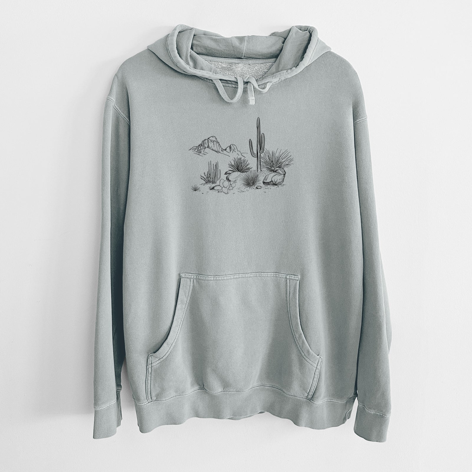 Sage green pigment dyed hoodie laid flat, printed with a black ink desert scene with a saguaro cactus, agave, rocks and distant mountains.