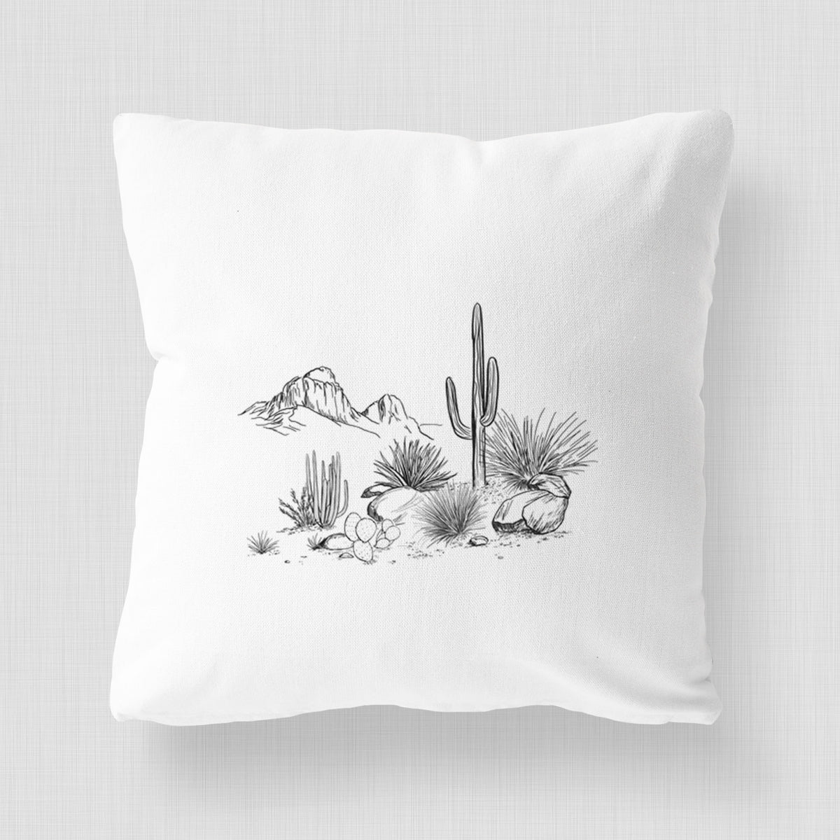 White pillow cover printed with a black ink desert scene with a saguaro cactus, agave, rocks and distant mountains.