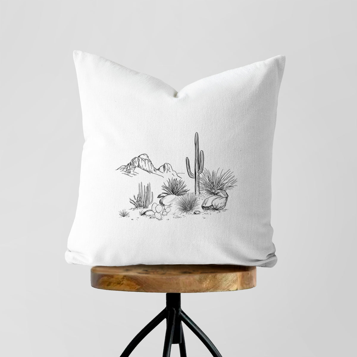 White pillow cover printed with a black ink desert scene with a saguaro cactus, agave, rocks and distant mountains, on a wooden stool.