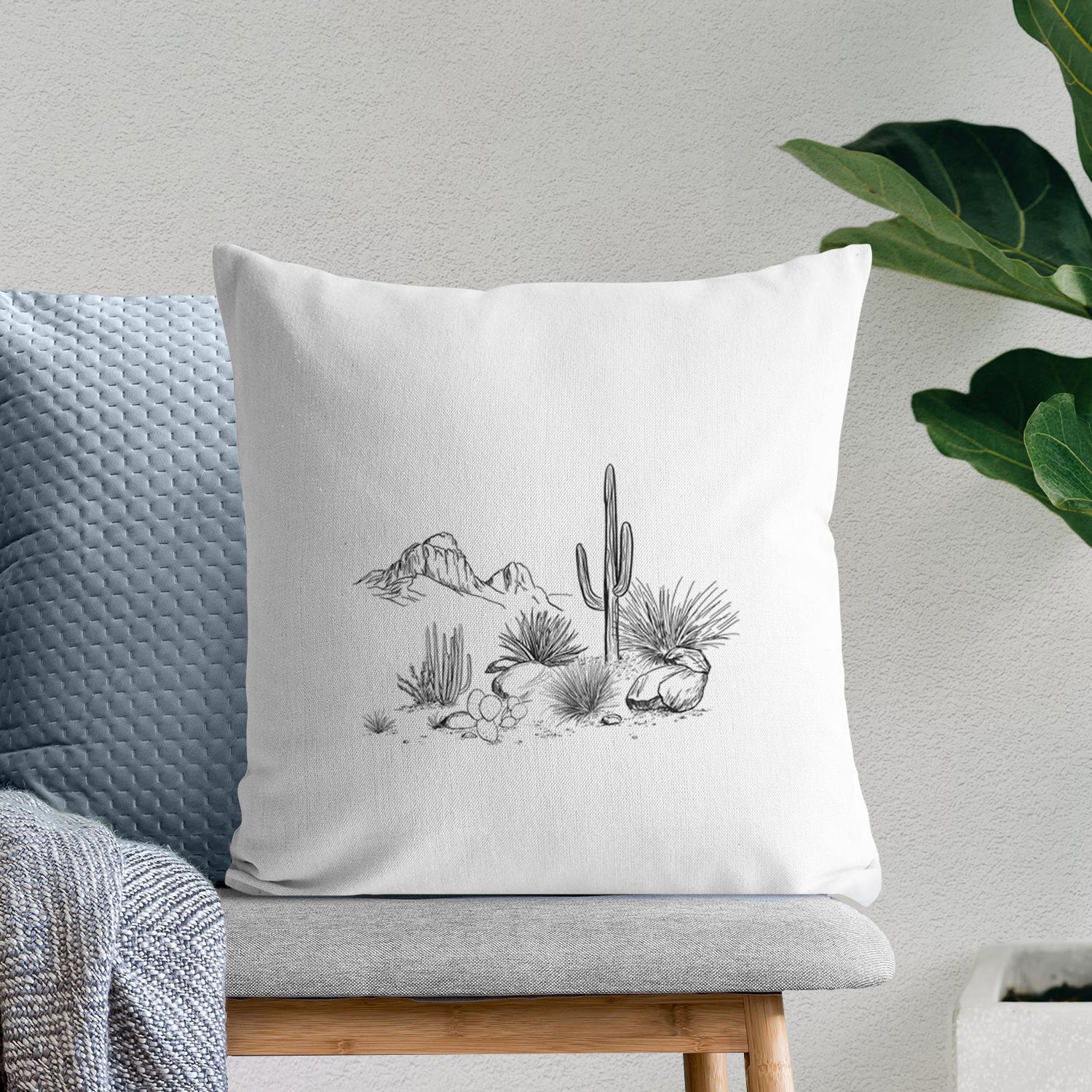White pillow cover printed with a black ink desert scene with a saguaro cactus, agave, rocks and distant mountains, on a bench beside plants.