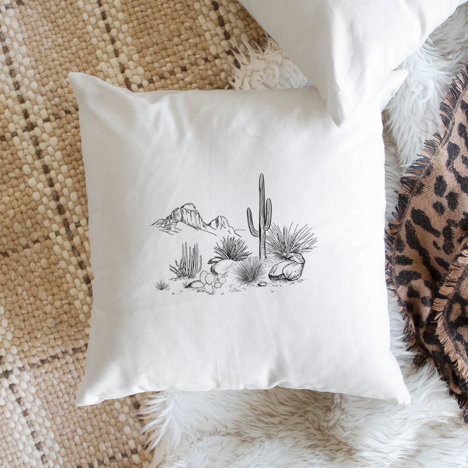 White pillow cover printed with a black ink desert scene with a saguaro cactus, agave, rocks and distant mountains, on a bench beside plants.