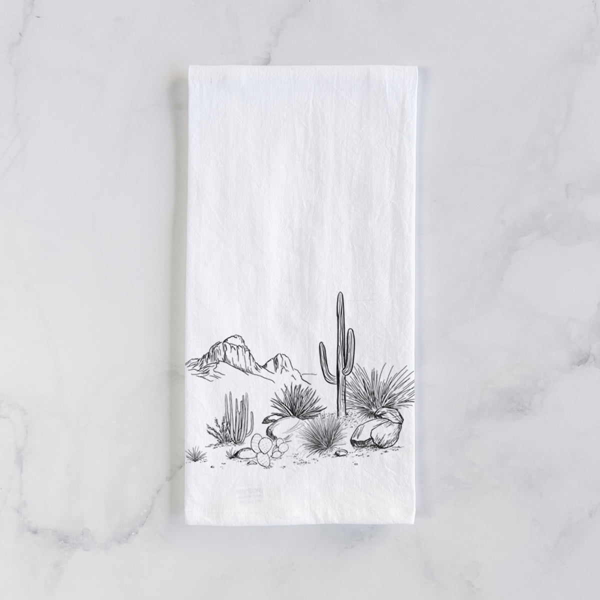 White cotton tea towel laid flat on marble, printed with a black ink desert scene with a saguaro cactus, agave, rocks and distant mountains.