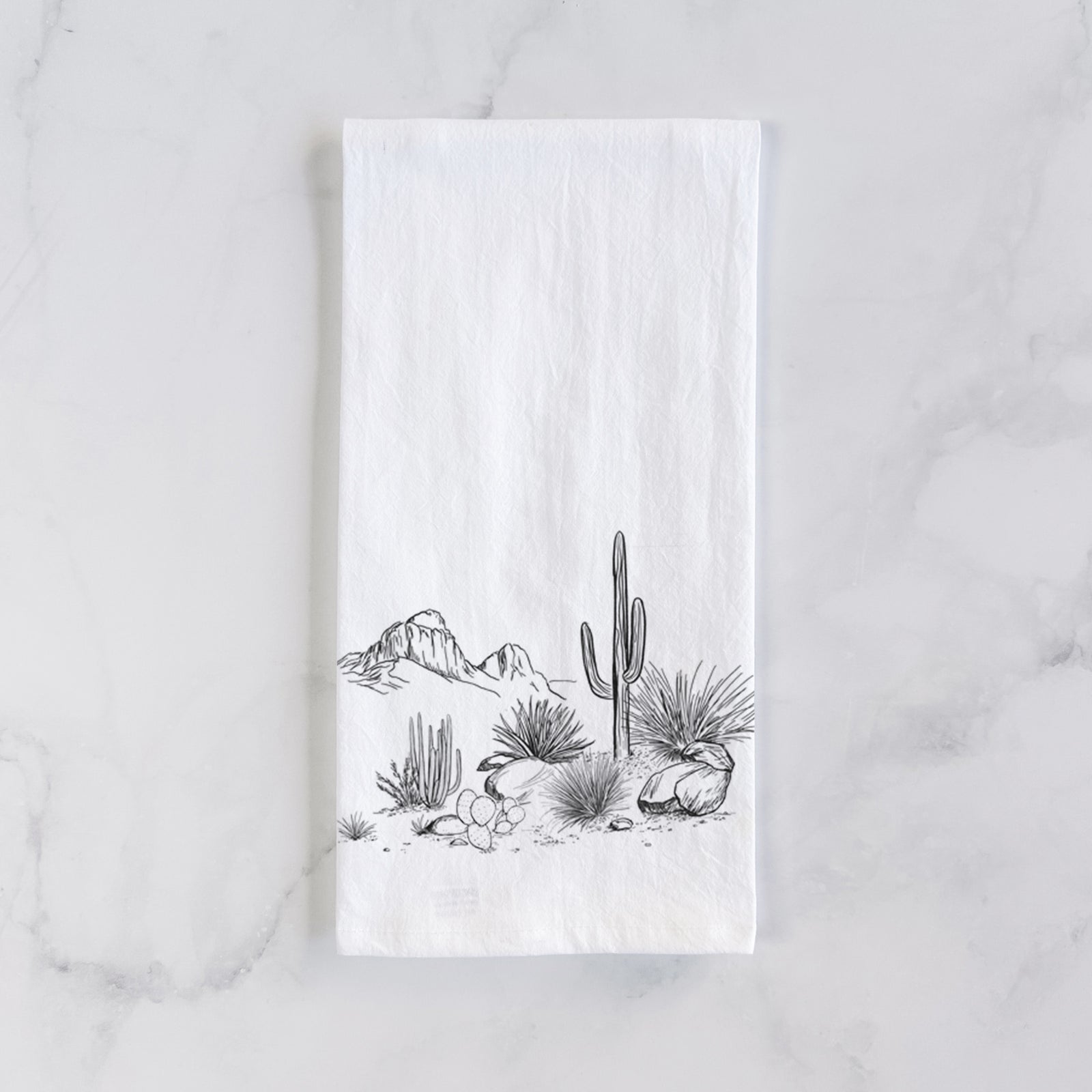 White cotton tea towel printed with a black ink desert scene with a saguaro cactus, agave, rocks and distant mountains, on a marble counter beside a round wooden cutting board and greenery.