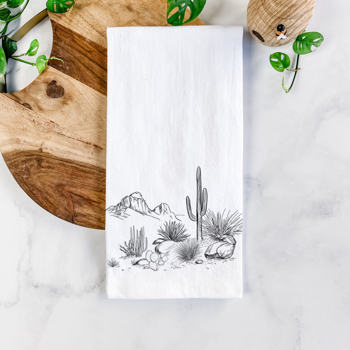 White cotton tea towel printed with a black ink desert scene with a saguaro cactus, agave, rocks and distant mountains, on a marble counter beside a round wooden cutting board and greenery.