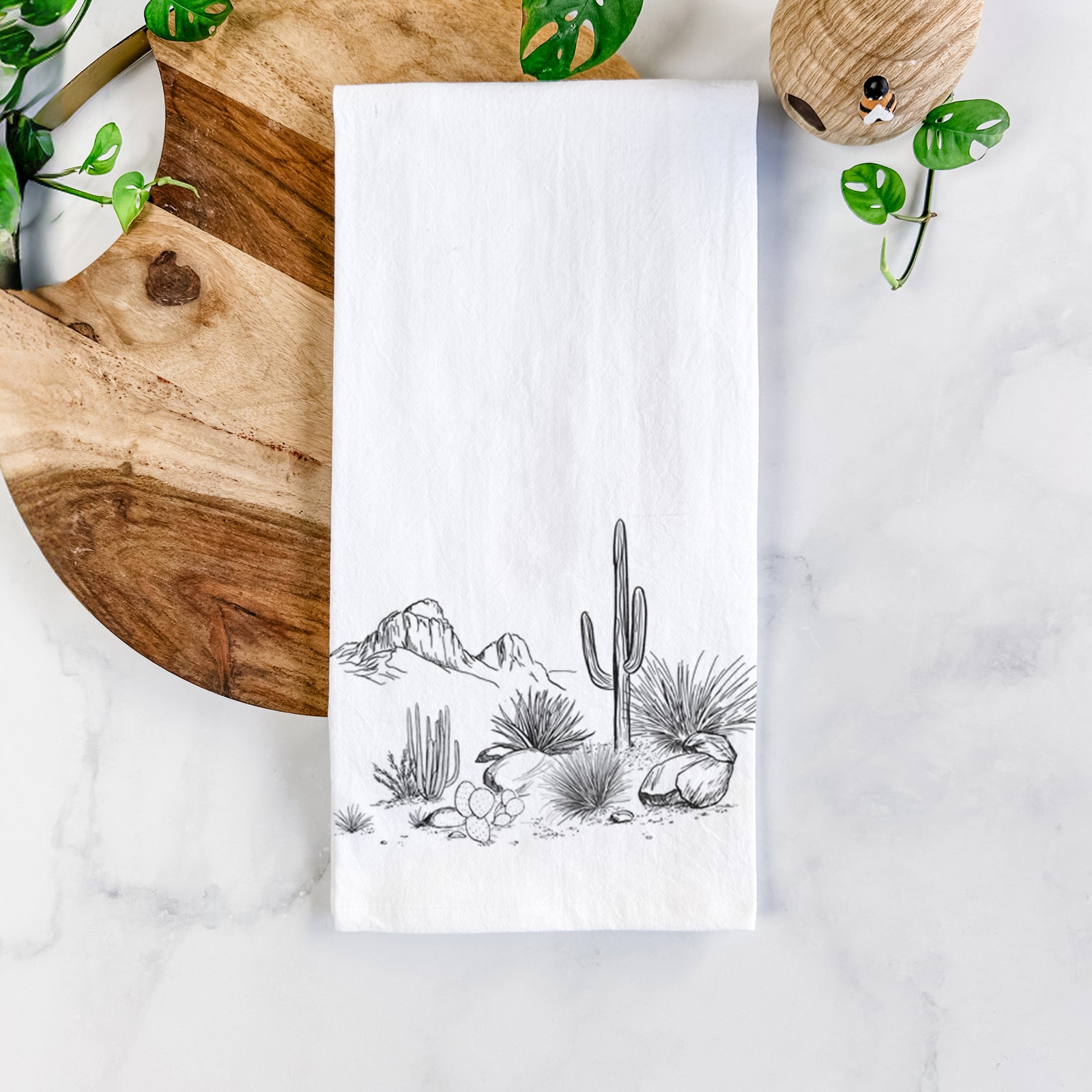 White cotton tea towel printed with a black ink desert scene with a saguaro cactus, agave, rocks and distant mountains, on a marble counter beside a round wooden cutting board and greenery.