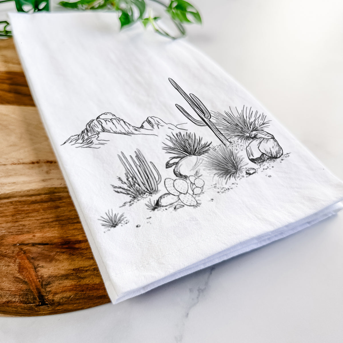 Close-up of a folded white tea towel on a wooden board, showing a black ink desert scene with a saguaro cactus, agave, rocks and distant mountains.