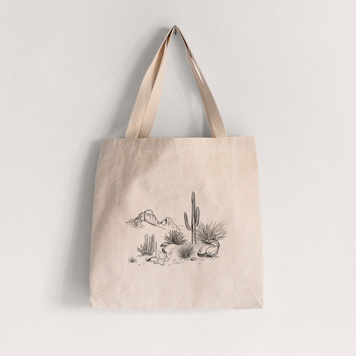 Natural canvas tote bag printed with a black ink desert scene with a saguaro cactus, agave, rocks and distant mountains.