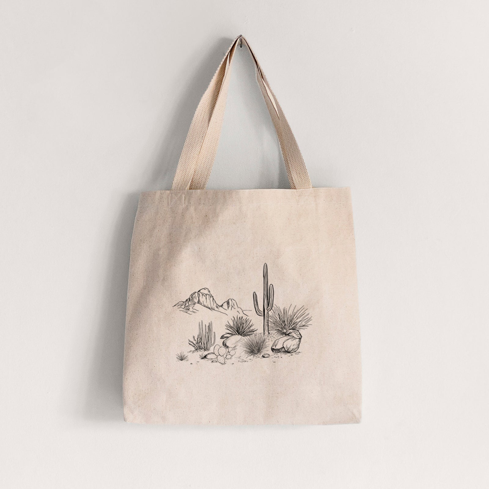 Natural canvas tote bag printed with a black ink desert scene with a saguaro cactus, agave, rocks and distant mountains.