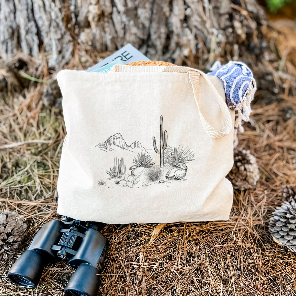 Canvas tote bag printed with a black ink desert scene with a saguaro cactus, agave, rocks and distant mountains, resting on dry grass beside a pair of binoculars.