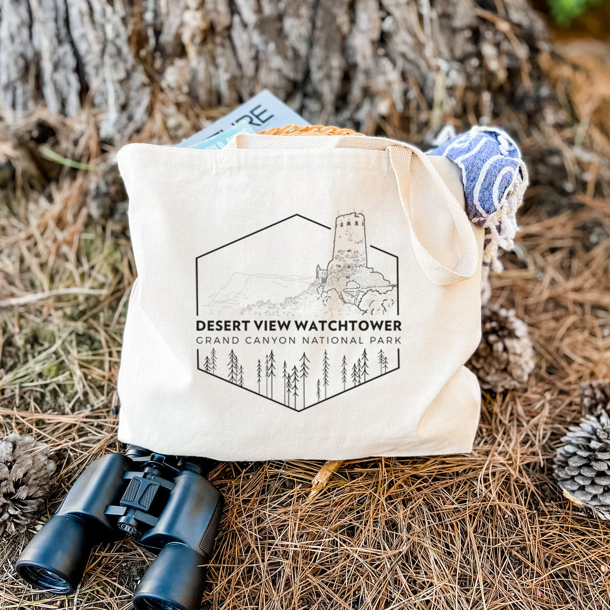 Canvas tote bag printed with a black line art badge of the Desert View Watchtower above pine trees, with the words Desert View Watchtower, Grand Canyon National Park, resting on dry grass beside a pair of binoculars.