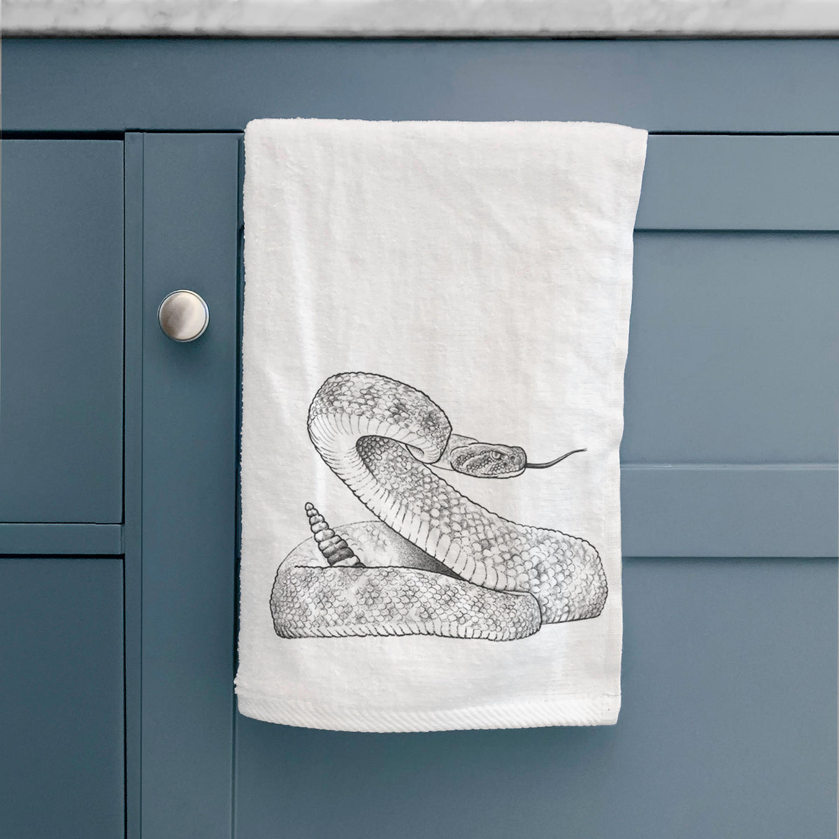 White hand towel printed with a detailed black ink illustration of a coiled diamondback rattlesnake, hanging on a cabinet door.